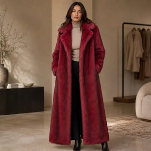 May include: A long, deep red faux fur coat with a wide collar and long sleeves. The coat is worn over a beige mock neck sweater and black trousers. The garment features a textured, shaggy pile finish and reaches down to the ankles.