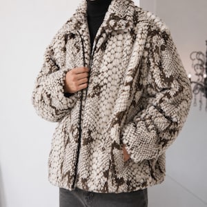 May include: A long-sleeved jacket featuring a textured, faux-fur material with a brown and cream snakeskin-inspired pattern. The jacket includes a fold-down collar, a full-length front zipper closure, and two side pockets, worn over a black turtleneck.