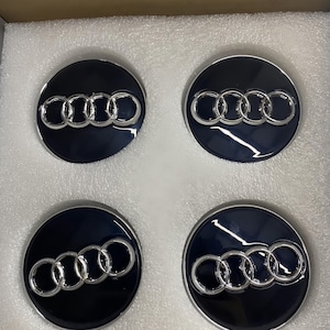 May include: Four black and silver car wheel center caps. Each cap features the Audi logo, a set of four interlocking rings, in silver against a black background. The caps are round and have a shiny, reflective surface.