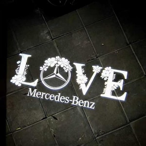 May include: A white illuminated projection on a dark tiled surface. The word "LOVE" is displayed with floral embellishments, incorporating the Mercedes-Benz logo. The text "Mercedes-Benz" is below.