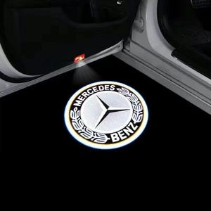Puede incluir: Un logotipo blanco y luminoso de Mercedes-Benz proyectado sobre una superficie negra cerca de una puerta de coche abierta. El logotipo circular muestra el nombre de la marca y una estrella de tres puntas. El interior del coche es visible.