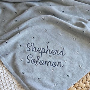 Puede incluir: Una manta de beb&eacute; de punto azul claro con el nombre "Shepherd Solomon" bordado. La manta tiene un delicado dise&ntilde;o calado y est&aacute; doblada sobre una cesta de mimbre. La manta es suave y acogedora.