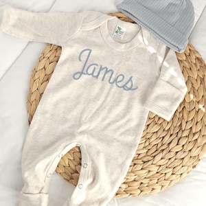 May include: A light beige baby onesie with the name "James" embroidered in blue script. A matching light blue ribbed knit cap sits beside the onesie. The items are displayed on a woven mat and white fabric.