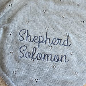 Puede incluir: Una manta de beb&eacute; azul claro con el nombre "Shepherd Solomon" bordado en hilo azul m&aacute;s oscuro. La manta tiene un delicado dise&ntilde;o calado con peque&ntilde;os agujeros espaciados uniformemente.