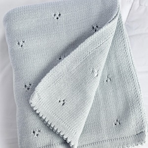 Puede incluir: Una manta de punto gris claro doblada, con un borde festoneado. La manta tiene un aspecto texturizado con peque&ntilde;os detalles de ojal espaciados uniformemente. El color suave y neutro y el dise&ntilde;o delicado sugieren una manta para beb&eacute; o un cubrecama decorativo.