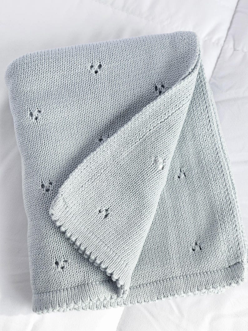 Puede incluir: Una manta de punto gris claro doblada, con un borde festoneado. La manta tiene un aspecto texturizado con peque&ntilde;os detalles de ojal espaciados uniformemente. El color suave y neutro y el dise&ntilde;o delicado sugieren una manta para beb&eacute; o un cubrecama decorativo.