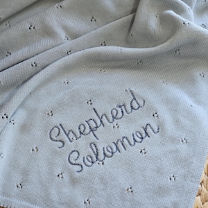 Puede incluir: Una manta de beb&eacute; de punto azul claro con el nombre "Shepherd Solomon" bordado. La manta presenta un delicado dise&ntilde;o calado y un borde festoneado. Se muestra sobre una superficie tejida.