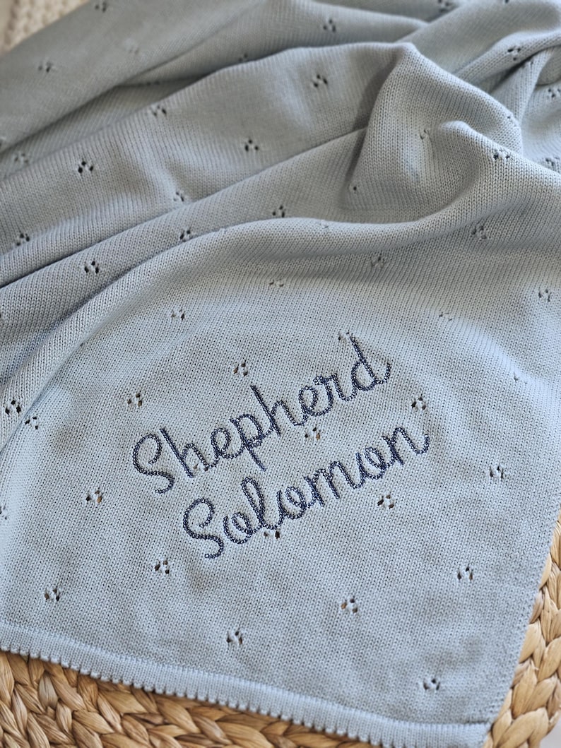 Puede incluir: Una manta de beb&eacute; de punto azul claro con el nombre "Shepherd Solomon" bordado. La manta presenta un delicado dise&ntilde;o calado y un borde festoneado. Se muestra sobre una superficie tejida.