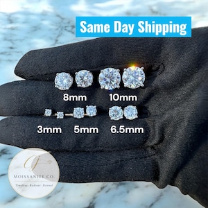 Puede incluir: Una selección de pendientes de moissanita de corte redondo en varios tamaños: 3 mm, 5 mm, 6,5 mm, 8 mm y 10 mm. Los pendientes se muestran sobre una superficie negra. El texto "Same Day Shipping" es visible.