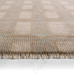 Puede incluir: Una alfombra anudada a mano con un patr&oacute;n de cuadr&iacute;cula repetitivo en tonos suaves de beige, crema y verde salvia apagado. La alfombra tiene una superficie texturizada de pelo y est&aacute; rematada con flecos cortos de color crema en el borde.