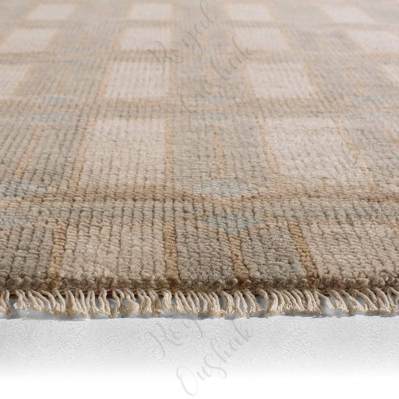 Puede incluir: Una alfombra anudada a mano con un patr&oacute;n de cuadr&iacute;cula repetitivo en tonos suaves de beige, crema y verde salvia apagado. La alfombra tiene una superficie texturizada de pelo y est&aacute; rematada con flecos cortos de color crema en el borde.