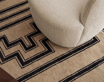 Puede incluir: Un sillón de bouclé beige con respaldo curvo y asiento redondeado, colocado sobre una alfombra rectangular de base beige con líneas geométricas negras audaces y patrones angulares que crean un diseño repetitivo tipo laberinto.