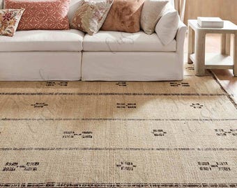 Puede incluir: Una alfombra beige tejida con patrones geométricos y rayas marrón oscuro sobre un suelo de madera. Un sofá blanco con cojines decorativos en tonos rosa, marrón y beige se encuentra encima de la alfombra.