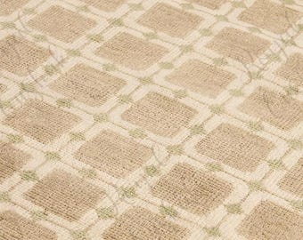 Puede incluir: Primer plano de una alfombra beige con un patrón geométrico repetitivo. El diseño presenta cuadrados en un color beige más oscuro, delineados con un beige más claro y acentuados con pequeñas formas de diamante verdes. La alfombra tiene una apariencia texturizada.