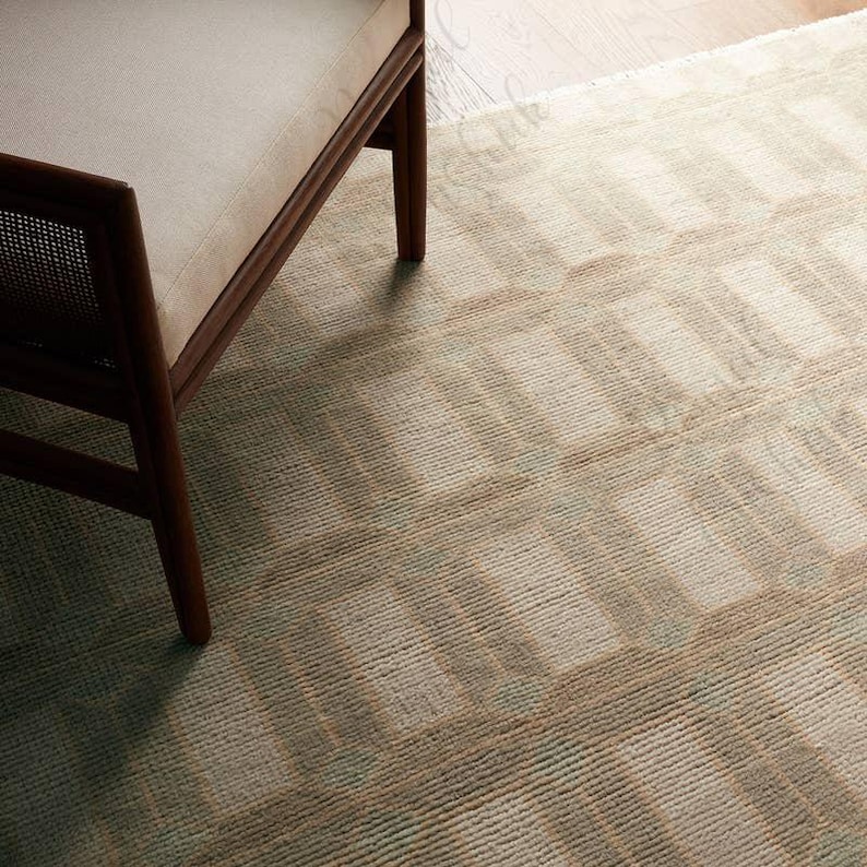 Puede incluir: Una alfombra tejida con un patr&oacute;n geom&eacute;trico repetitivo de bloques rectangulares en tonos apagados de beige, crema y verde salvia suave. La alfombra tiene una superficie texturizada de pelo corto y est&aacute; parcialmente cubierta por la pata y el marco de madera de una silla.