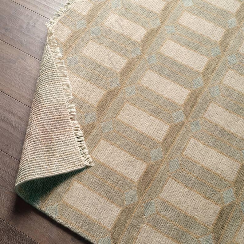 Puede incluir: Una alfombra tejida con un patr&oacute;n geom&eacute;trico repetitivo de rect&aacute;ngulos de color beige claro delineados en verde salvia. La alfombra tiene una superficie texturizada y un borde con flecos donde la esquina est&aacute; doblada hacia atr&aacute;s.