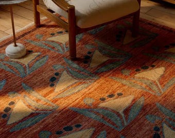 Puede incluir: Una alfombra rectangular con un estampado floral en tonos naranja, amarillo y azul. La alfombra está colocada sobre un suelo de madera, con una silla de madera y una mesa auxiliar en el fondo. El diseño de la alfombra presenta flores y hojas estilizadas.
