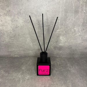 May include: A black reed diffuser with a square base and a bright pink label featuring the text Furz loscher in black lettering. Three thin black reeds extend upwards from the centre of the bottle, which has a matte finish and a minimalist design.