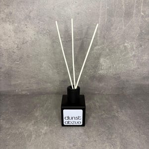 Könnte beinhalten: Ein quadratischer Diffusor aus schwarzem Glas mit drei weißen Stäbchen, die oben eingesteckt sind. Auf der Vorderseite befindet sich ein weißes quadratisches Etikett mit dem schwarzen Schriftzug "dunst abzug" in einer modernen serifenlosen Schrift.