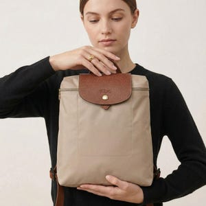Peut inclure: Un sac à dos beige avec un rabat et une poignée en cuir marron. Le sac à dos a une fermeture éclair et un petit bouton doré sur le rabat. Les bretelles et le dos du sac à dos sont également marron.