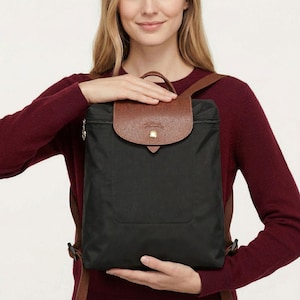 May include: Black backpack with a brown leather flap and handle. The backpack has brown shoulder straps and a gold-coloured zip. The flap is secured with a gold-coloured press stud. The backpack is made of a durable, water-resistant material.