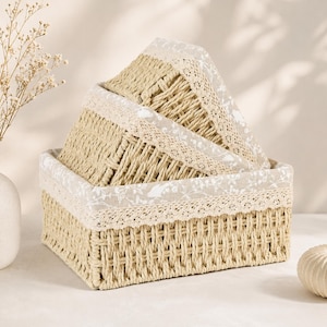 May include: A set of three rectangular woven baskets made of light tan natural fibres. Each basket features a beige fabric liner with a white floral pattern and a decorative cream-coloured lace trim along the top edge. The baskets are nested together.