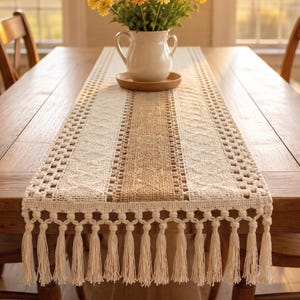 Pode incluir: Caminho de mesa bege e castanho claro com design tecido e detalhes de franjas. O caminho de mesa está centrado numa mesa de madeira, com um vaso de flores amarelas ao fundo.