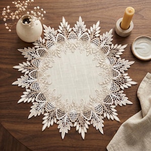 Puede incluir: Un tapete circular color crema con intrincados detalles de encaje. El tapete presenta un tejido central similar al lino rodeado por un borde de encaje detallado, de inspiración floral. El diseño está sobre una superficie de madera oscura, con un pequeño jarrón, una vela y un plato cerca.