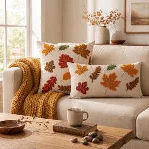 Puede incluir: Dos cojines decorativos de color crema con hojas de otoño bordadas en tonos rojo, naranja, amarillo, marrón y verde. Un cojín es cuadrado y el otro rectangular, ambos muestran un patrón disperso de varias formas de hojas.