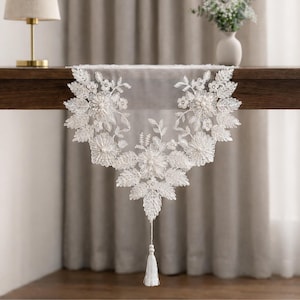 Può includere: Runner da tavolo in pizzo bianco con ricami floreali e a foglie. Il runner presenta un design delicato con dettagli di perline e un nappa all'estremità. È esposto su un tavolo di legno scuro.