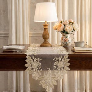 May include: A wooden table is set with a decorative runner, lamp, and floral arrangement. The runner is beige with an embroidered floral design. A wooden lamp with a white shade sits on the table. A vase of flowers and a teacup are also on the table.