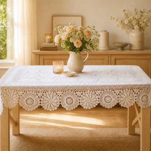 May include: A white, rectangular crochet tablecloth with a scalloped edge featuring a floral design. The tablecloth is draped over a light-colored wooden table. A vase of flowers and a lit candle are on the table.