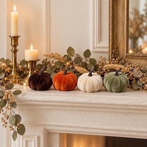 Puede incluir: Cuatro calabazas de terciopelo en marrón oscuro, naranja, crema y verde salvia, cada una con un tallo marrón oscuro. Están dispuestas en fila junto a dos candelabros de latón con velas blancas encendidas y una guirnalda de eucalipto y trigo seco.