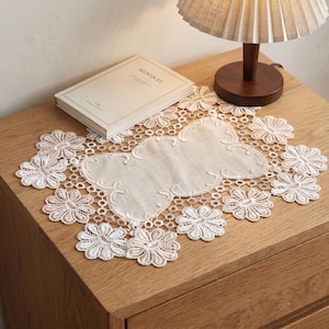 May include: A white lace doily with a floral design, featuring a central oval shape and surrounding flower motifs. The doily is placed on a wooden surface, alongside a book and a lamp.