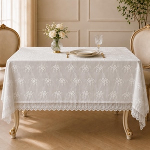 Puede incluir: Un mantel rectangular blanco con un intrincado patrón de bordado floral en la superficie y un borde decorativo de encaje festoneado. La tela cubre una mesa, la cual está decorada con un florero de cristal con flores blancas.