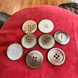 May include: A collection of eight round buttons in various colors and designs. The buttons are made of materials like mother-of-pearl and feature two or four holes for sewing. Some buttons have decorative carvings.