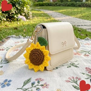 May include: A cream-colored shoulder bag with a flap closure and a small embroidered bow detail. A crocheted sunflower charm with a brown center, yellow petals, and a green leaf is attached to the bag's gold-toned metal hardware ring.