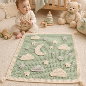 May include: A rectangular crochet baby blanket featuring a sage green center with a cream-colored border and corner tassels. The blanket is decorated with appliqued cream and grey crochet clouds, stars, and a crescent moon across the surface.