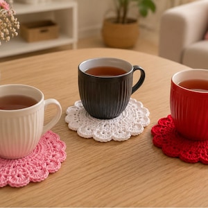 Puede incluir: Tres tazas de cerámica con texturas acanaladas verticales, llenas de té, sobre posavasos redondos de ganchillo. Las tazas son blancas, negras y rojas, mientras que los posavasos a juego son rosas, blancos y rojos, sobre una superficie de madera.