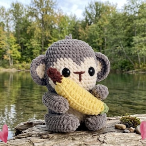 May include: A handmade crochet monkey doll in shades of gray and cream, holding a yellow banana. The monkey has large black eyes and a brown mouth. The doll is sitting on a piece of driftwood near a body of water.