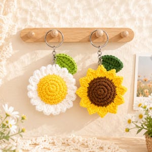 May include: Two crochet flower keychains hanging from a wooden rack. One keychain features a white daisy with a yellow center and a green leaf. The other keychain is a yellow sunflower with a dark brown center and a green leaf. Both have silver rings.