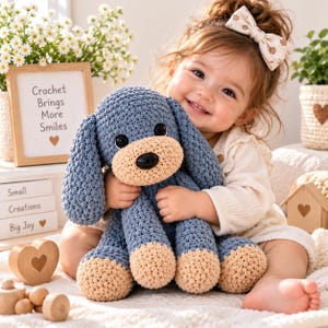 May include: A blue and beige crocheted dog plush toy with large black plastic eyes and a black nose. The toy features a textured yarn construction with floppy ears and rounded paws, designed with a soft, handmade aesthetic for tactile play.