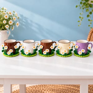 Puede incluir: Un juego de cinco tazas de cerámica en marrón, blanco, marrón oscuro, crema y morado, cada una sobre un posavasos de ganchillo verde a juego decorado con margaritas blancas y amarillas que se extienden por los lados de las tazas.