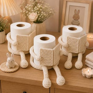 May include: Three cream-colored crochet toilet paper holders, each designed to look like a figure sitting and holding an open book. The holders are made of textured yarn and feature rounded arms and legs that wrap around standard toilet paper rolls.