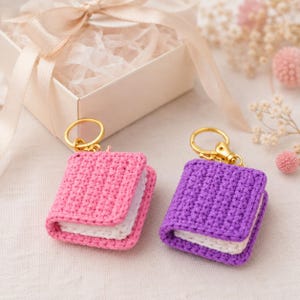 Pode incluir: Dois porta-chaves de livro em miniatura feitos de fio de croché. Um livro tem uma capa cor-de-rosa brilhante, enquanto o outro tem uma capa roxa. Ambos os artigos incluem páginas brancas de fio e estão presos a argolas de metal dourado.