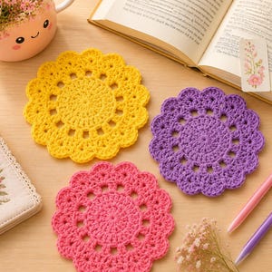 May include: Three round, crocheted coasters in bright yellow, purple, and pink, each featuring a scalloped edge and a central circular pattern. The coasters are arranged on a light-coloured surface alongside a book, a pink mug, and two pens.