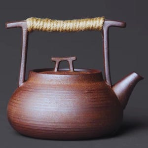 May include: A brown ceramic teapot with a rope handle. The teapot has a rounded body, a spout, and a lid with a small handle. The handle is made of a natural-coloured rope wrapped around a brown metal frame.