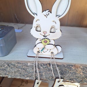 Könnte beinhalten: Holzhasenfigur mit langen Ohren, die eine gelbe Blume hält. Der Hase hat hängende Beine aus Holz und Schnur. Die Figur sitzt auf einem Holzsockel, perfekt für die Osterdekoration.