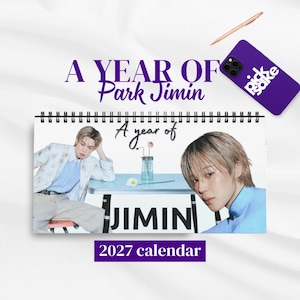May include: A 2027 spiral-bound calendar titled A YEAR OF Park Jimin. The cover features two photographs of a person with light hair, a light blue table with a small vase and flower, and the text A year of JIMIN. A purple phone case is nearby.