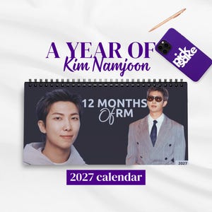 May include: A 2027 spiral-bound wall calendar titled A YEAR OF Kim Namjoon and 12 MONTHS Of RM. The cover features two portraits of Kim Namjoon, one in a hoodie and one in a suit with sunglasses. A purple phone case and a pen are nearby.